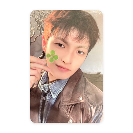 ATEEZ Hongjoong Blue Drift event photocard — reddish-brown hair, brown fringed suede jacket over a denim shirt, four-leaf clover pressed to his cheek, open field and bare tree branches behind him.