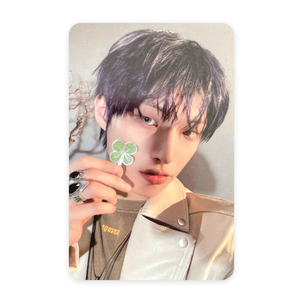 ATEEZ Mingi Blue Drift event photocard — dark violet-black hair, cream leather jacket with lettering, chunky silver rings, holding a four-leaf clover at his face, bare branches behind him.