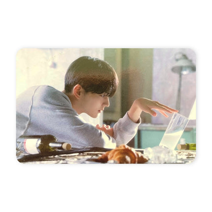 ENHYPEN Jungwon THE SIN : BLISS Music Korea POB landscape photocard — dark hair, oversized gray sweatshirt, leaning across a cluttered table and reaching toward a glass of milky drink beside a wine bottle, croissant and scattered ice, teal wall and vintage wall lamp behind him.